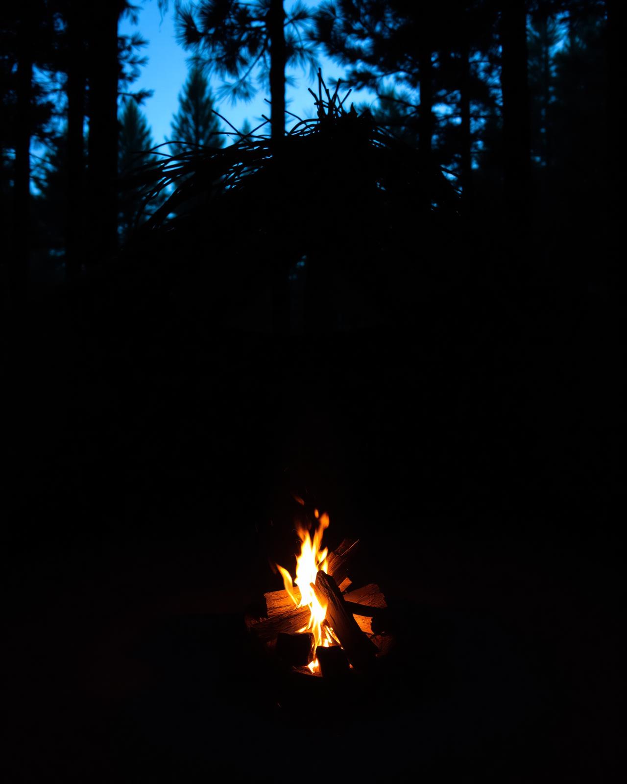 A small campfire glowing in front of a hand-built shelter at dusk